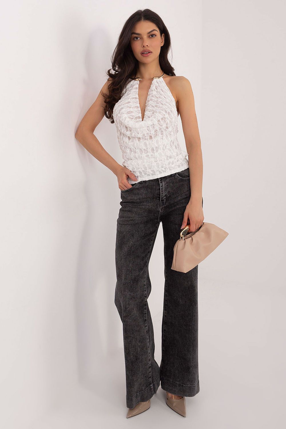 Woman wearing a white sleeveless top and dark jeans, holding a beige clutch against a plain background