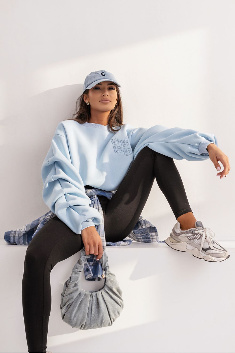 Woman sitting on a white surface wearing a light blue sweatshirt, black leggings, and gray sneakers.
