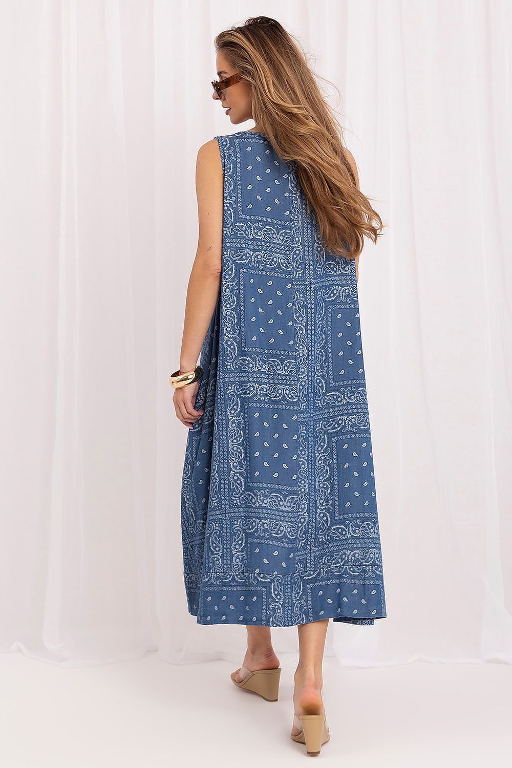 A woman wearing a sleeveless blue midi dress with a white bandana print pattern, shown from the back against a white curtain backdrop.