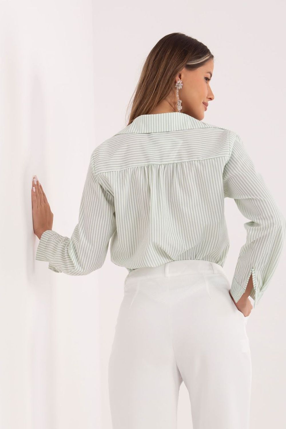 A woman shown from the back wearing a light sage green and white vertically striped long-sleeve blouse tucked into white trousers, posing against a clean white wall.