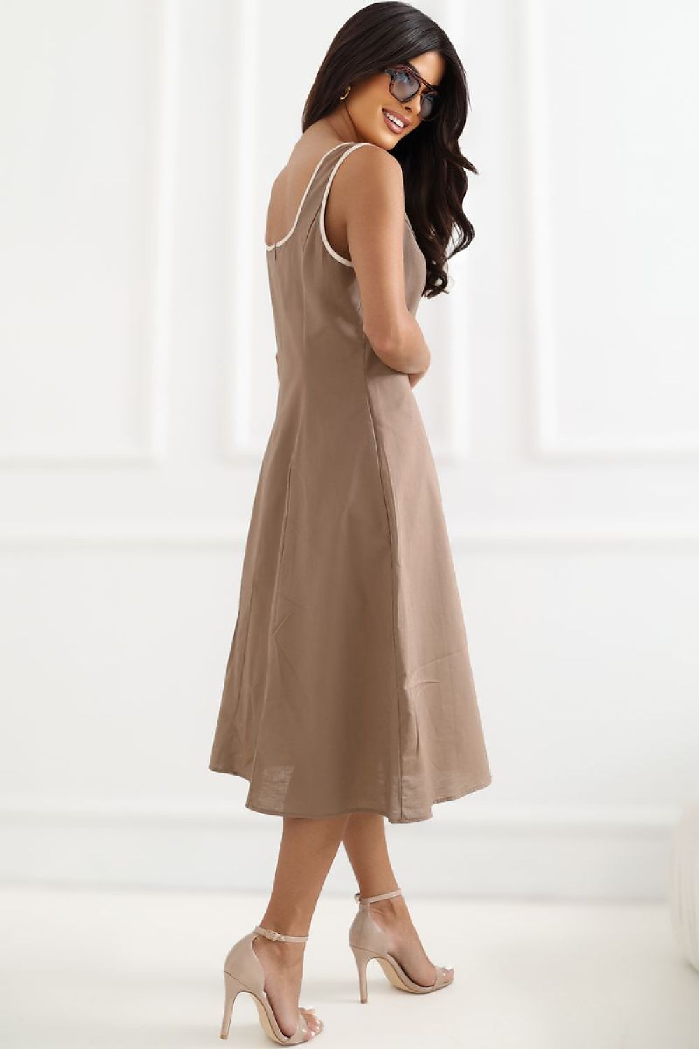 A woman wearing a sleeveless taupe midi dress with thin white trim along the straps and neckline, paired with beige high-heeled sandals, photographed in a bright white interior setting.