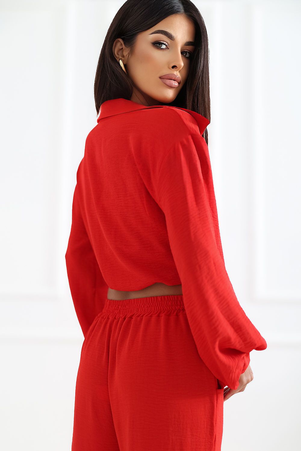 A woman wearing a bright red two-piece outfit, featuring a cropped long-sleeve top with a high collar and matching wide-leg trousers, shown from a three-quarters back view against a white paneled background.