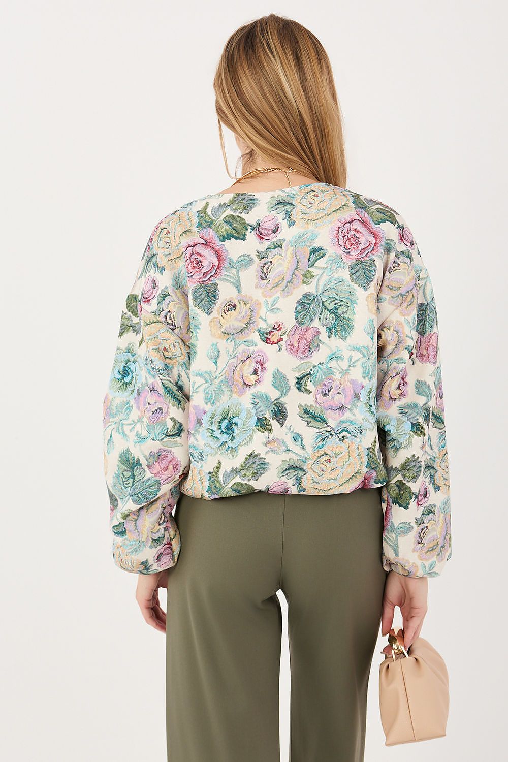 A woman shown from the back wearing a cropped, long-sleeve floral jacket with a cream base and pink, green, and blue floral pattern, paired with olive green trousers and holding a small beige handbag against a plain white background.