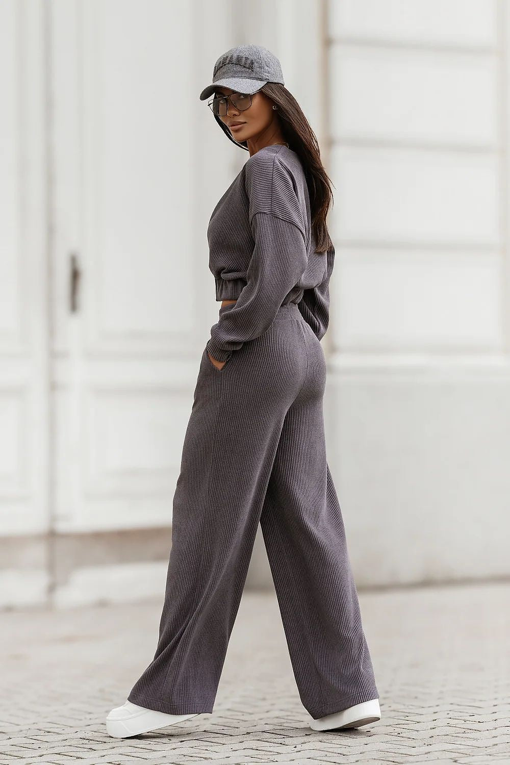 A woman wearing a matching dark gray lounge set consisting of a cropped long-sleeve top and wide-leg pants, paired with a gray baseball cap and white sneakers, walking outdoors against a white building background.
