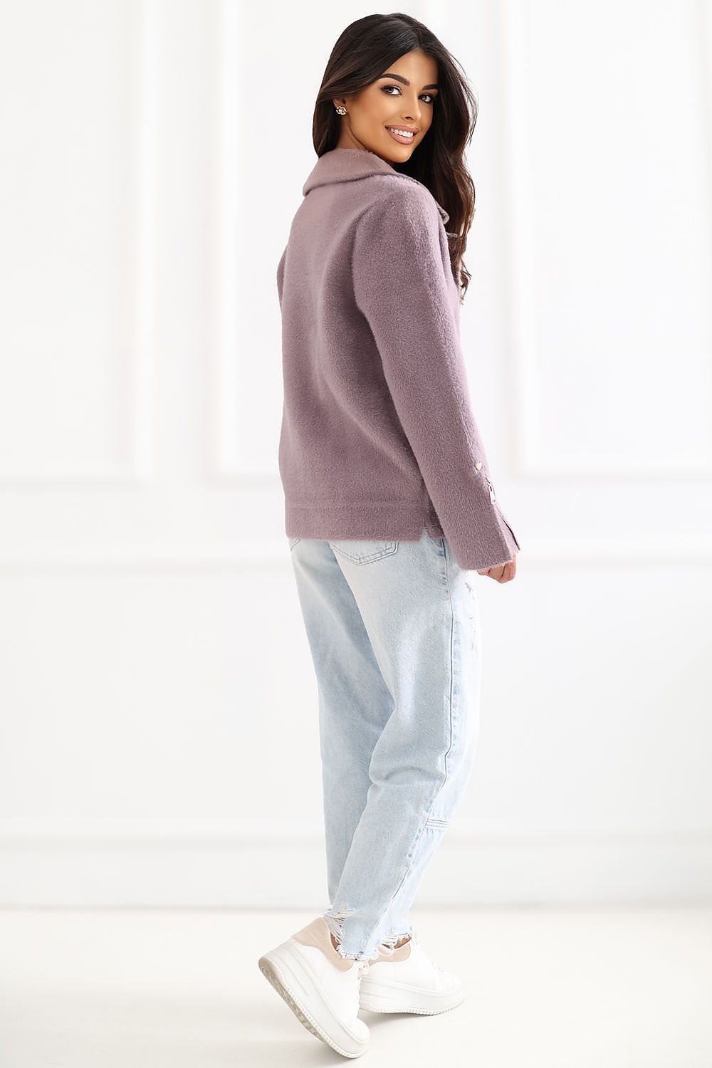A woman modeling a dusty mauve knit cardigan sweater, paired with light-wash jeans and white sneakers, shown from a side-back angle against a bright white background.