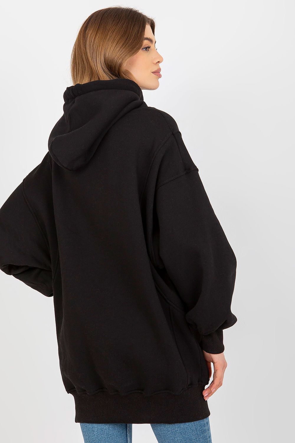 A woman wearing an oversized black pullover hoodie, shown from a side-back angle against a plain light gray studio background.