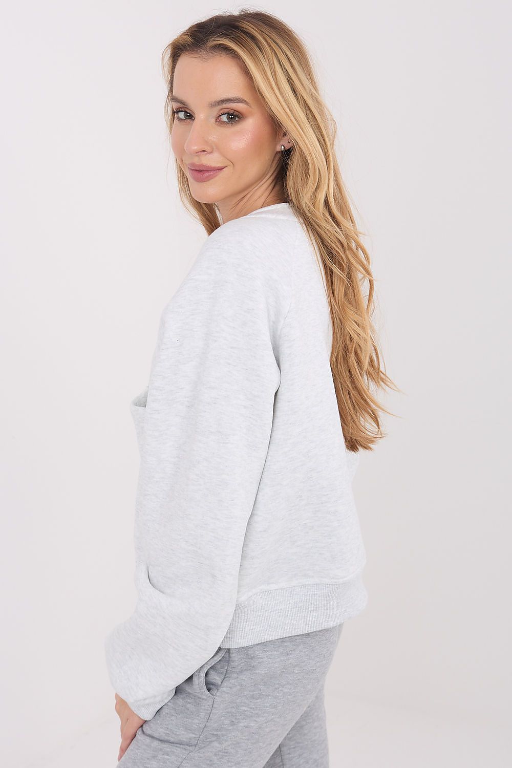 A woman with blonde hair wearing a light gray heather crewneck sweatshirt and gray sweatpants, shown from a side-back angle against a plain white studio background.