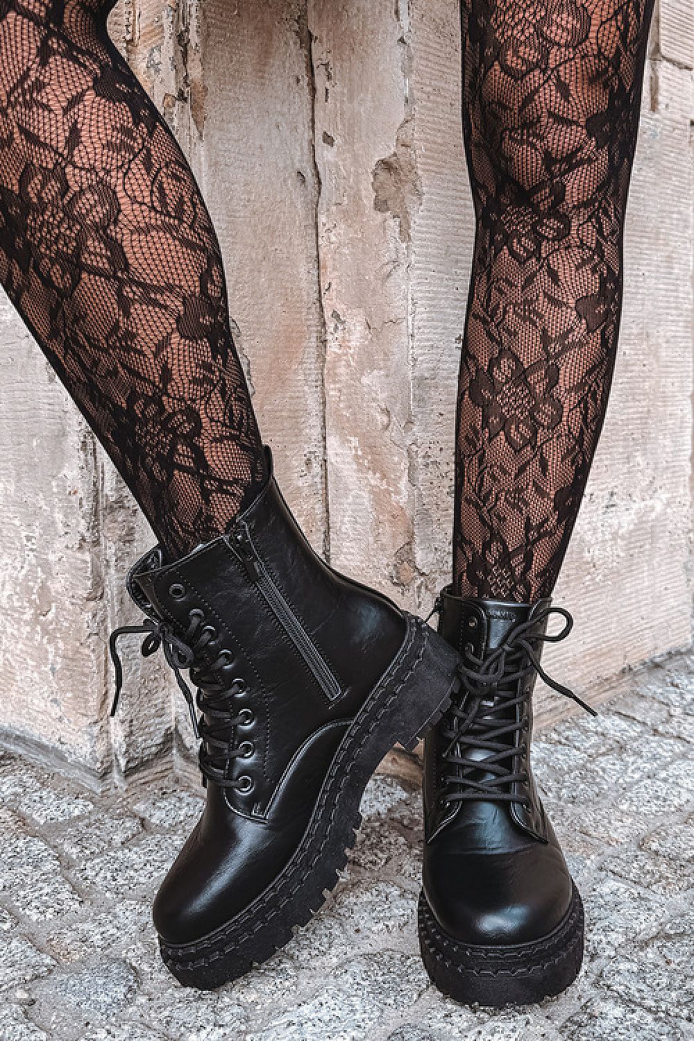 Black lace-up ankle boots with side zippers and chunky lug soles, shown on a model wearing black floral lace tights against a rustic stone wall.