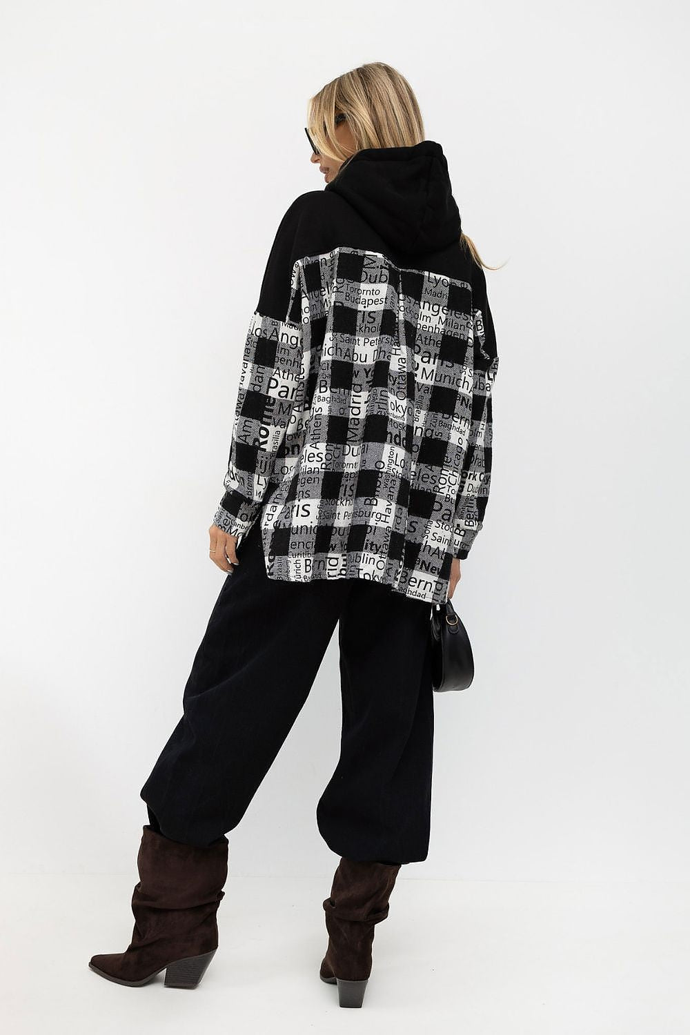 A woman shown from the back wearing a black and white graphic plaid hoodie with a solid black hood, paired with black wide-leg pants and dark brown heeled boots, against a plain white studio background.