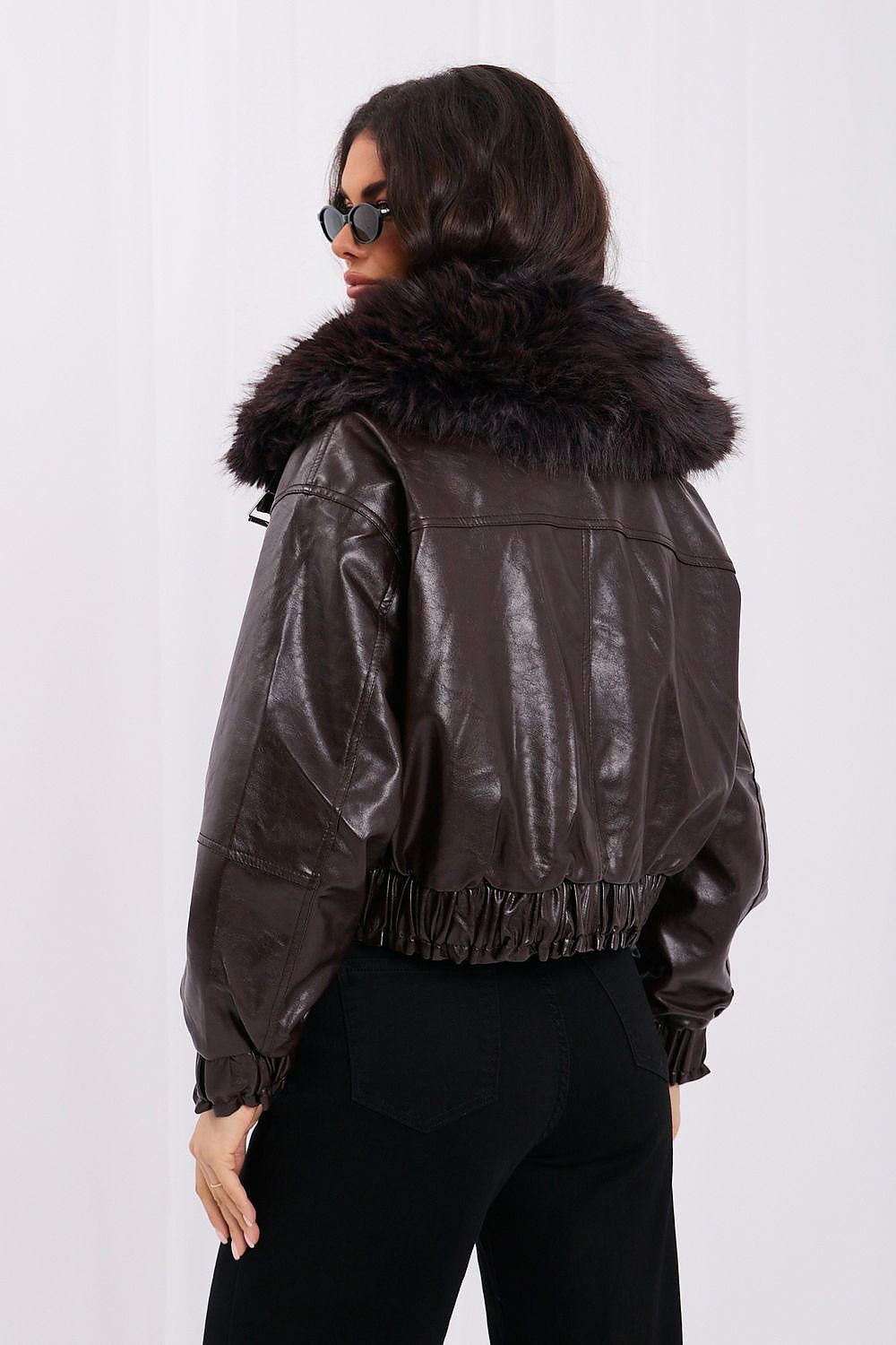 A woman shown from the back wearing a dark brown faux-leather jacket with a large dark brown faux-fur collar, paired with black pants, against a white studio background.
