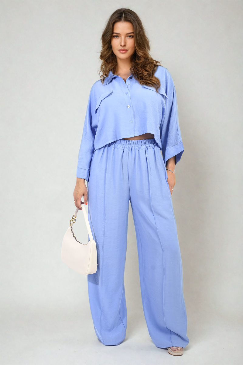 Woman wearing a light blue outfit with wide-leg pants and a matching top, holding a white handbag on a plain background.