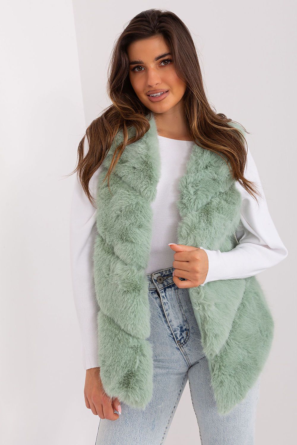 Woman wearing a light green fluffy vest over a white shirt and jeans on a white background