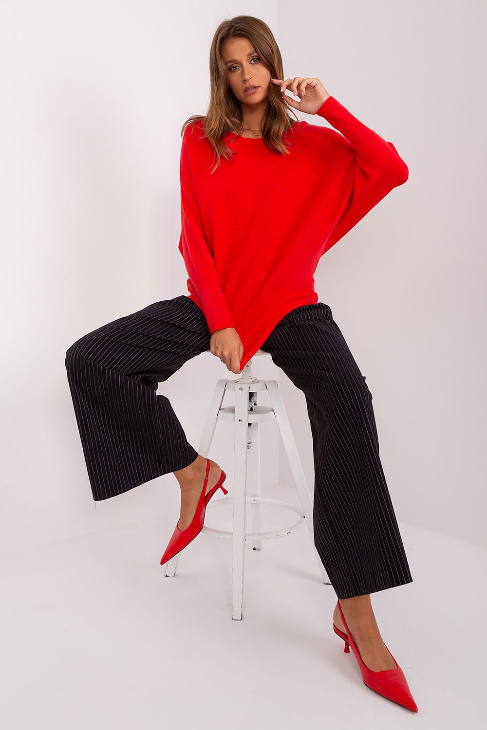 Woman in a red sweater and black pants sitting on a white stool against a white background