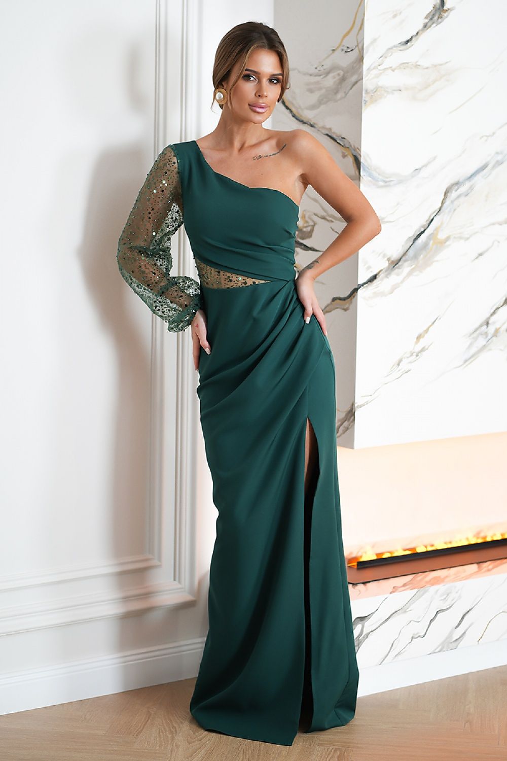 Woman wearing a green one-shoulder evening gown with a slit, standing against a marble wall.