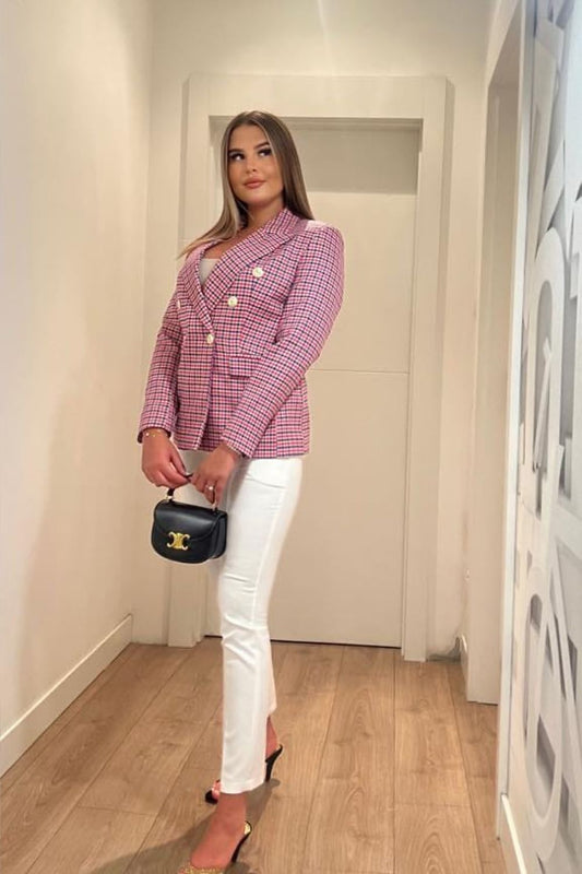Woman wearing a pink checkered blazer and white pants, holding a black handbag in a hallway.