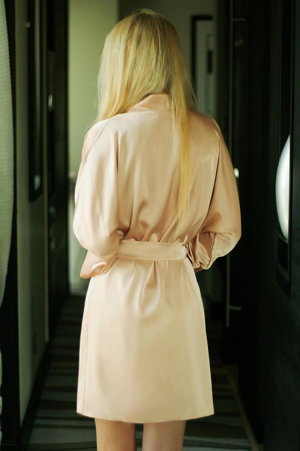 Woman wearing a beige trench coat in a dimly lit hallway.