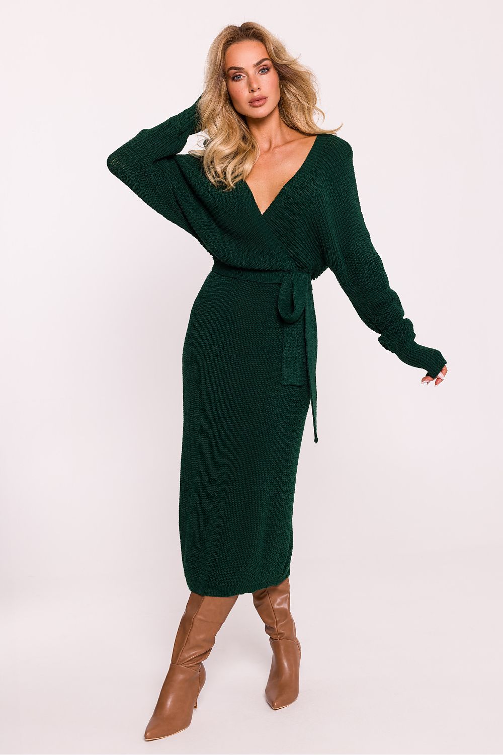 A woman modeling a dark forest green knit wrap midi dress with long sleeves, a V-neckline, and a matching tie belt, paired with brown knee-high boots against a plain light background.