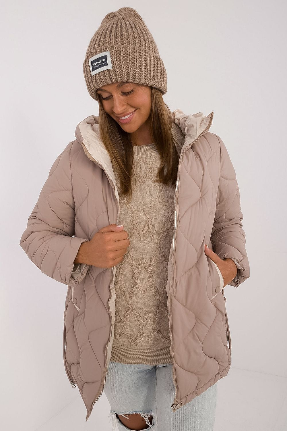 Woman wearing a beige quilted jacket, knit hat, and sweater on a white background