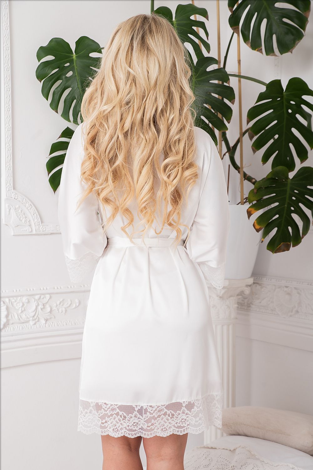 Person wearing a white lace-trimmed dress with long, wavy blonde hair in a room with green plants.