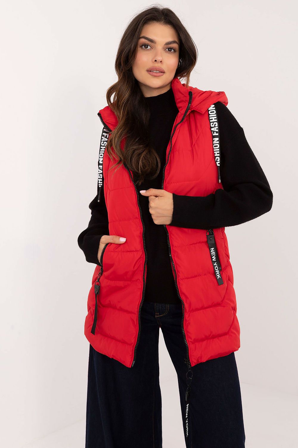 A woman modeling a bright red puffer vest with black trim and white text details on the shoulder straps, worn over a black long-sleeve top and dark pants against a plain light background.