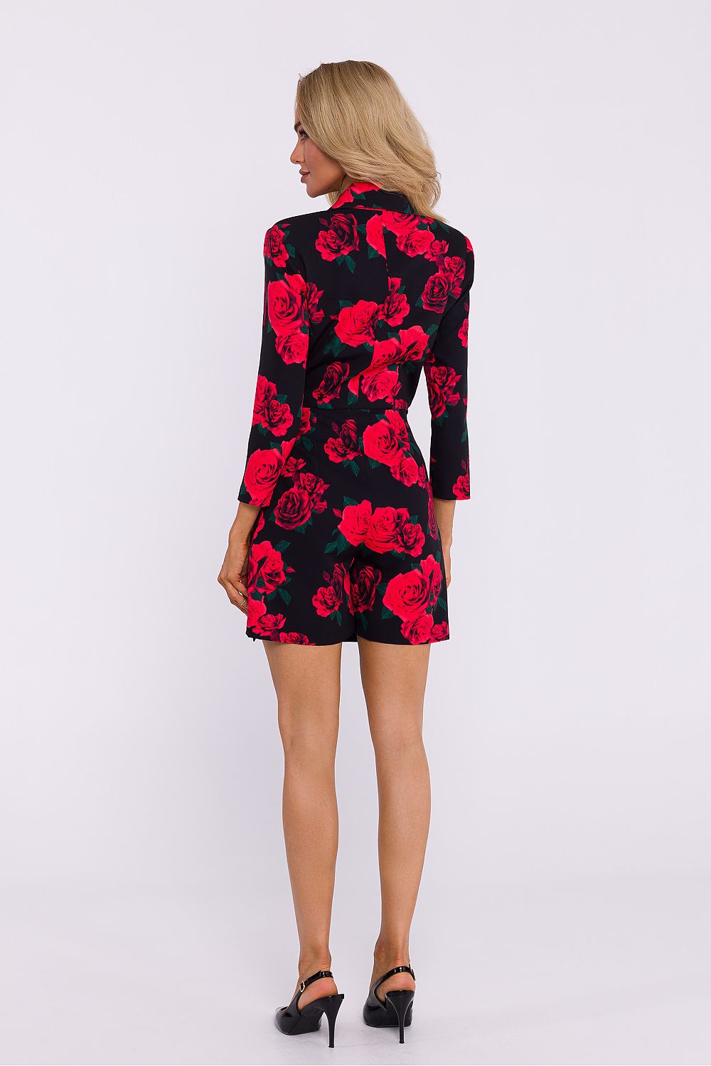 A woman viewed from the back wearing a black long-sleeve mini dress with a vibrant red rose pattern, paired with black high heels, against a plain white studio background.