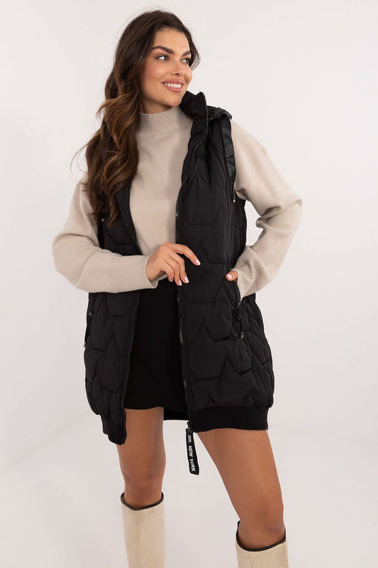 Woman wearing a black puffer vest over a beige sweater and skirt on a white background