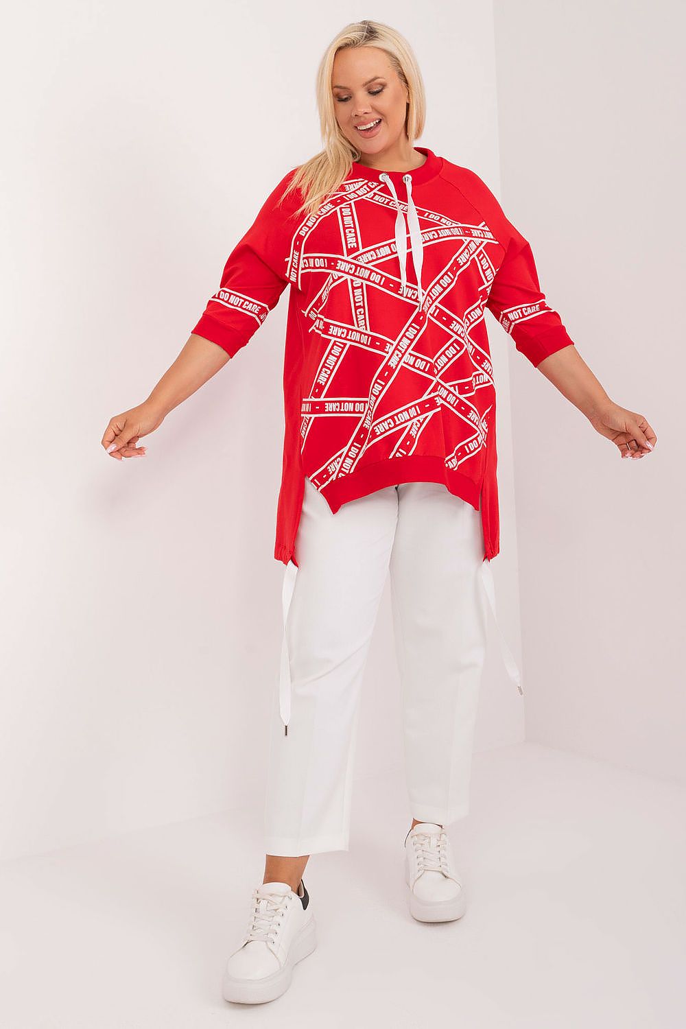 Woman wearing a red patterned top and white pants on a plain background