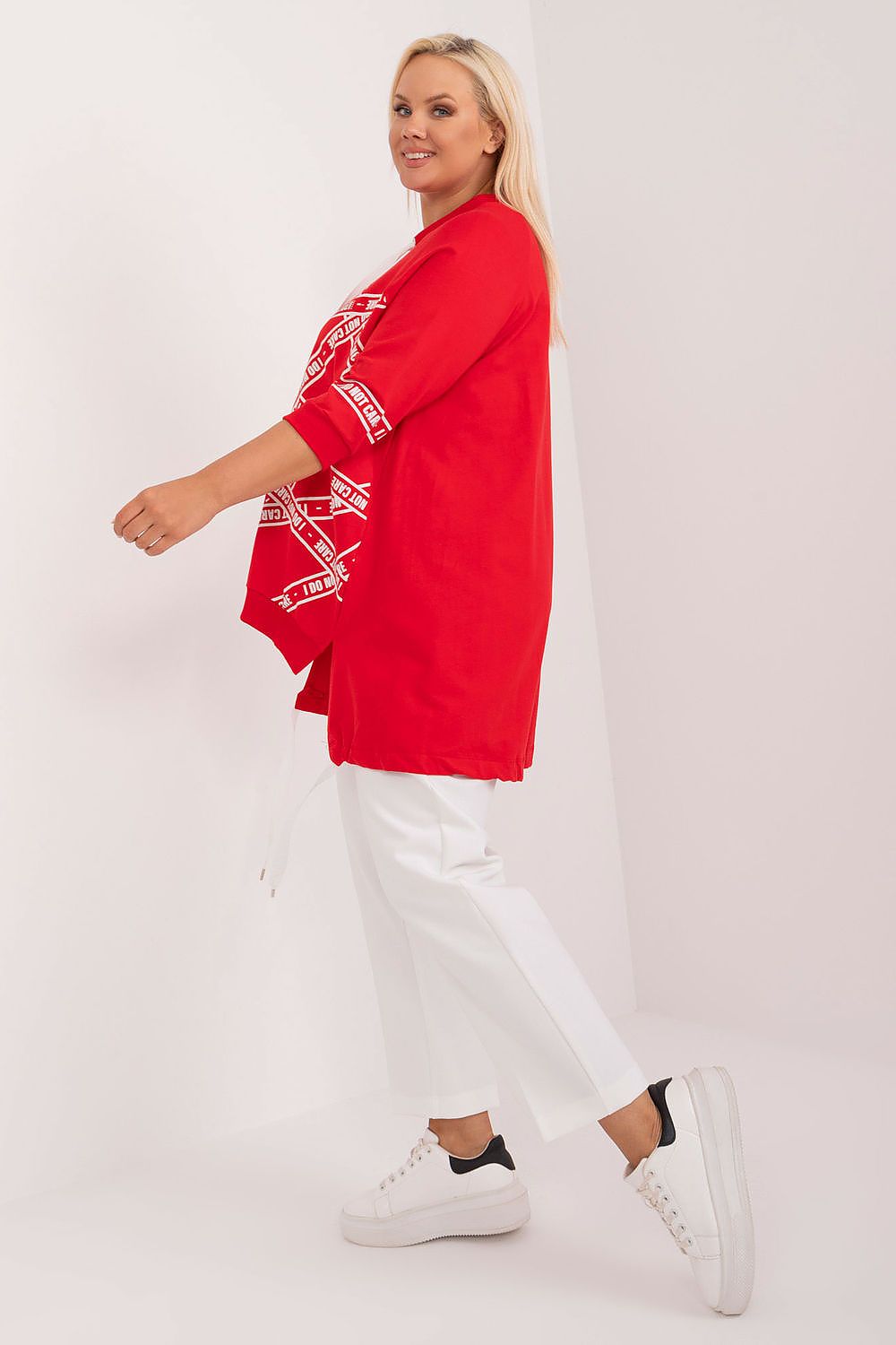 Woman wearing a red cardigan with white patterns and white pants on a plain background