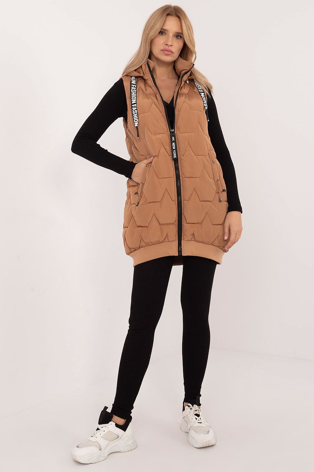 Woman wearing a tan quilted vest over a black long-sleeve shirt on a white background
