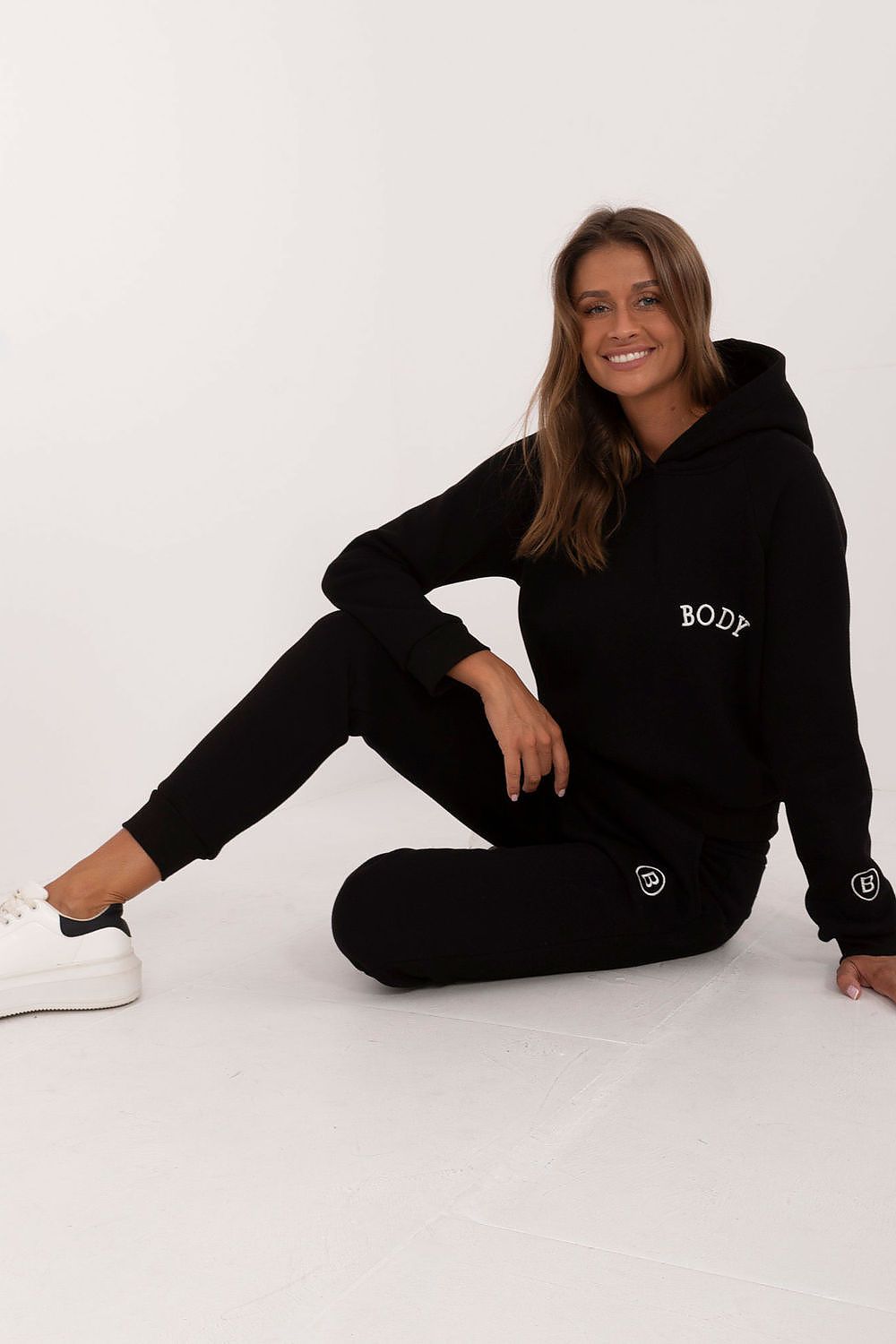 Woman wearing a black hoodie and pants sitting on a white floor.