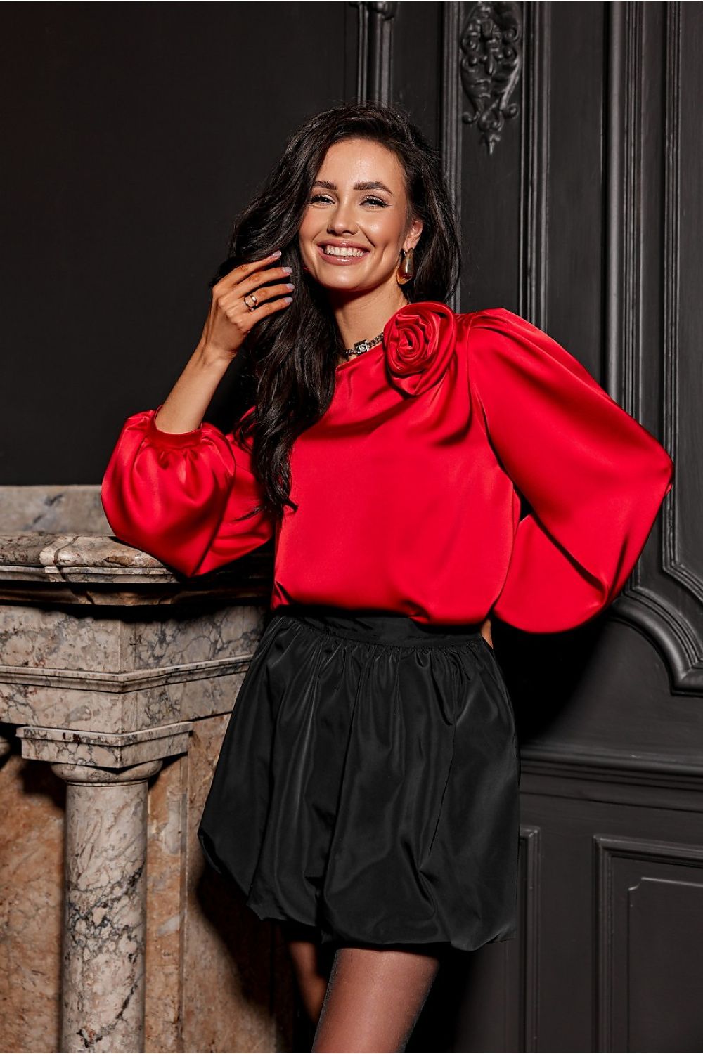 Woman in a red blouse and black skirt standing against a dark wall.