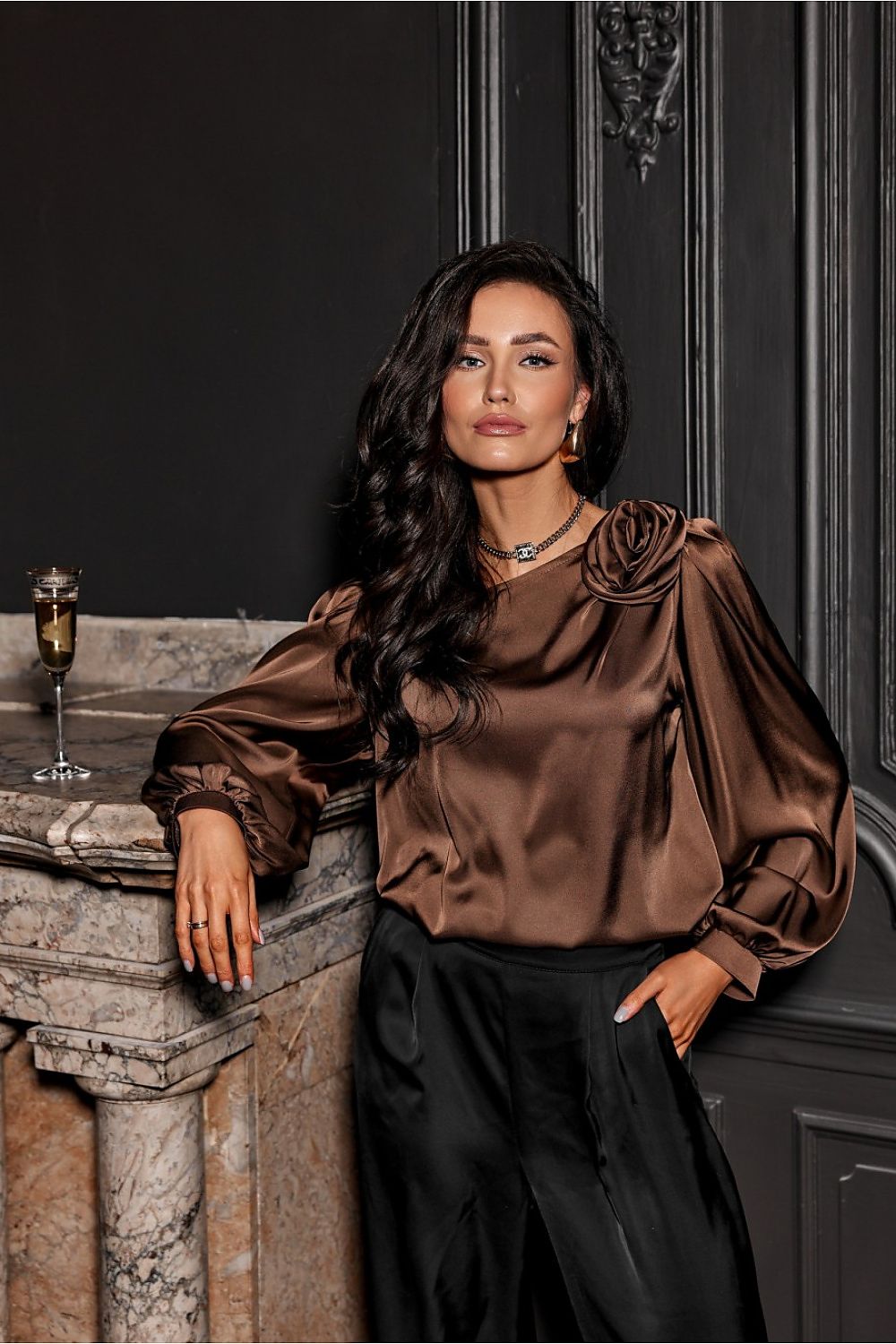 Woman in a brown satin blouse leaning against a stone wall with a dark background