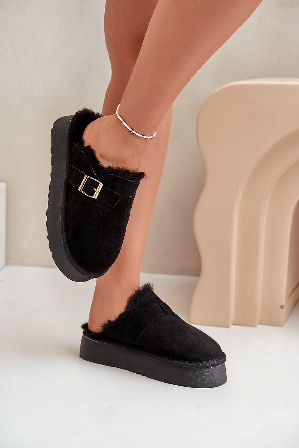 Black slippers with fur trim worn by a person on a neutral background