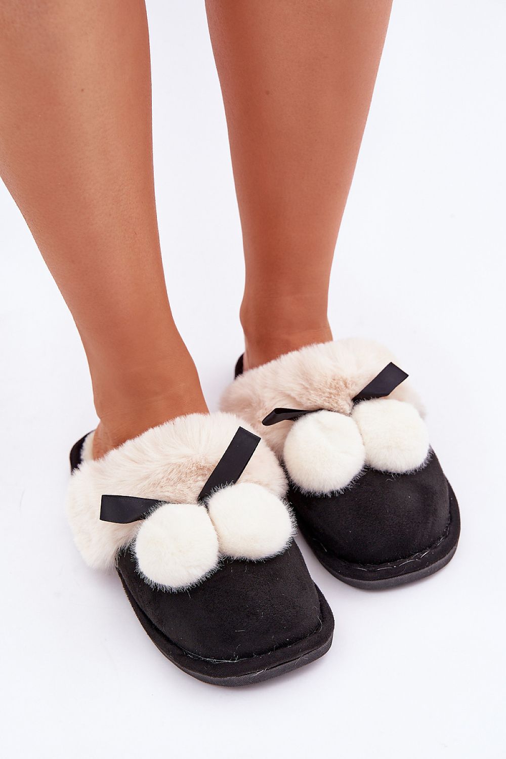 Black and white slippers with pom-poms and bows worn by a person on a white background