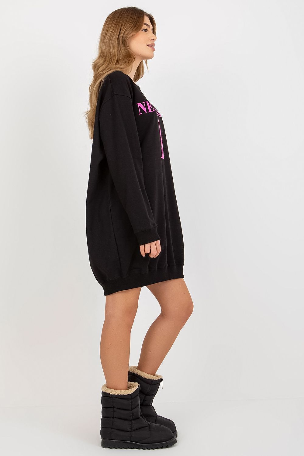 Person wearing a black oversized sweatshirt with pink text and black boots on a white background