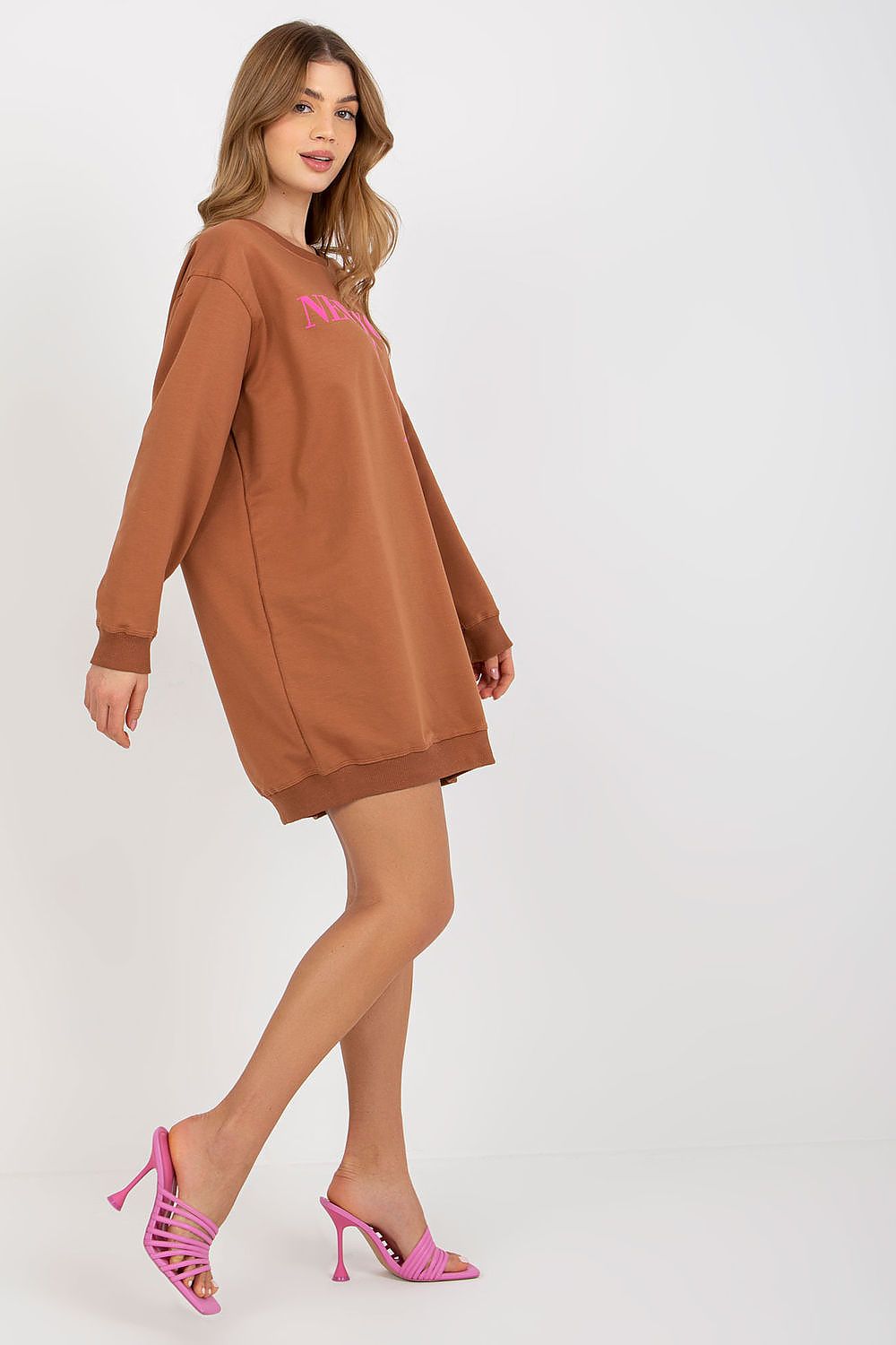 Woman wearing a brown sweatshirt dress with pink text and pink high-heeled shoes on a white background