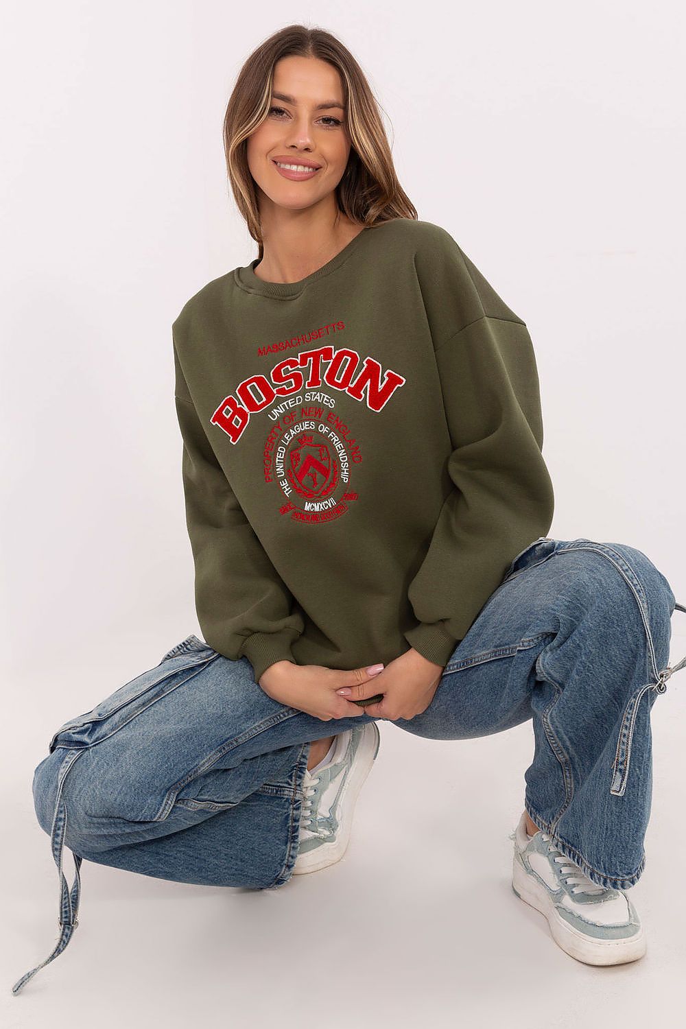 Woman wearing a green sweatshirt with 'Boston' print, sitting on the floor against a white background