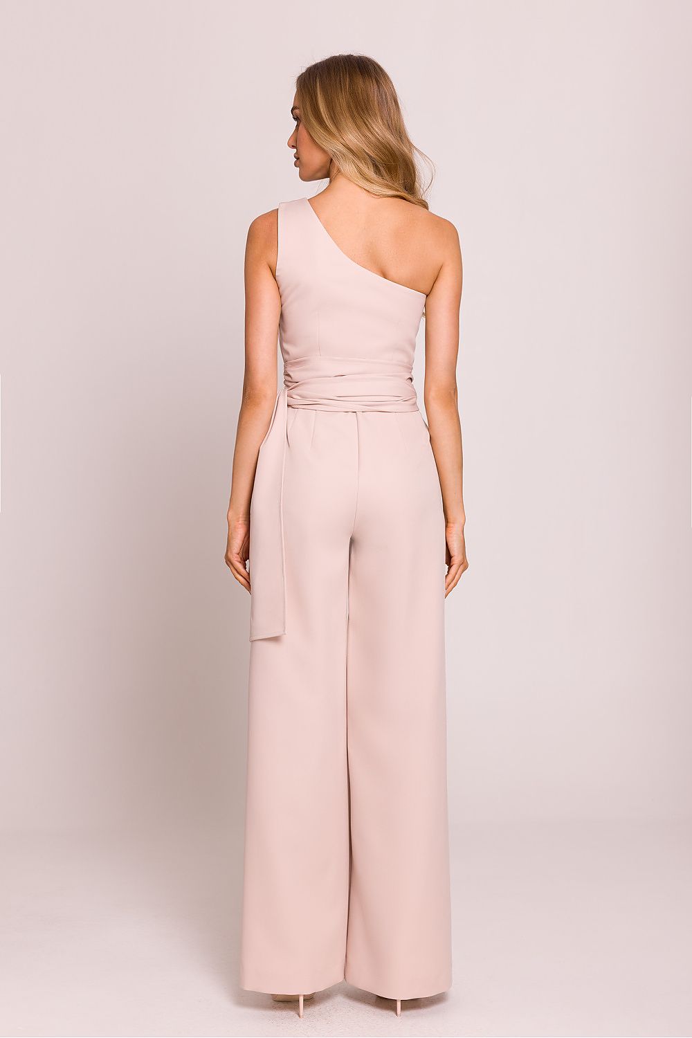 Woman wearing a light pink one-shoulder jumpsuit on a plain background
