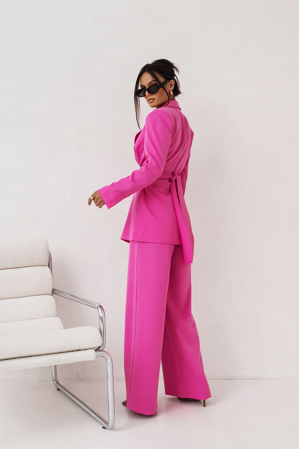 Person wearing a bright pink suit standing in a minimalistic room with a white chair.
