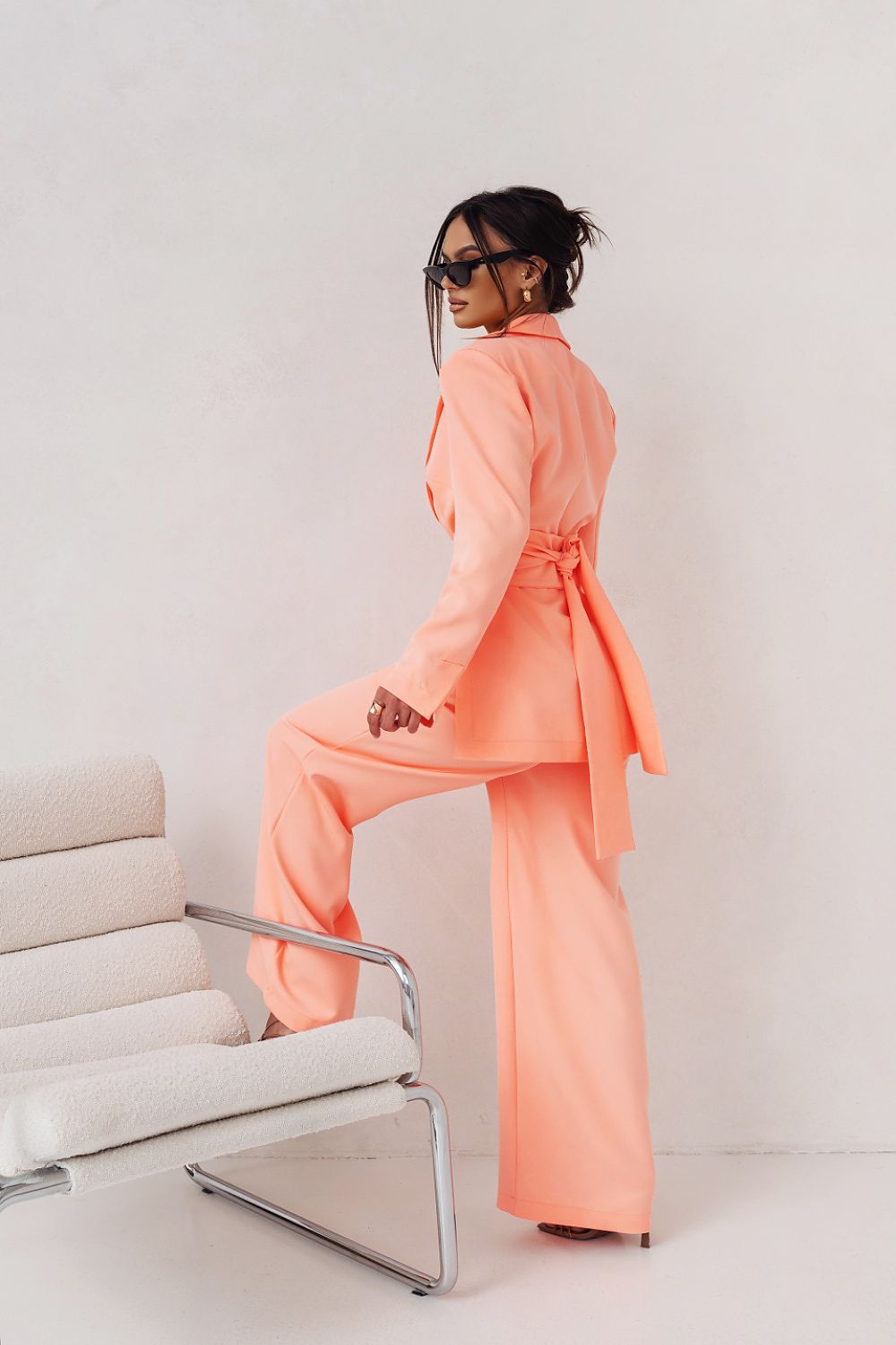 Woman in a peach-colored outfit standing next to a white chair against a plain background