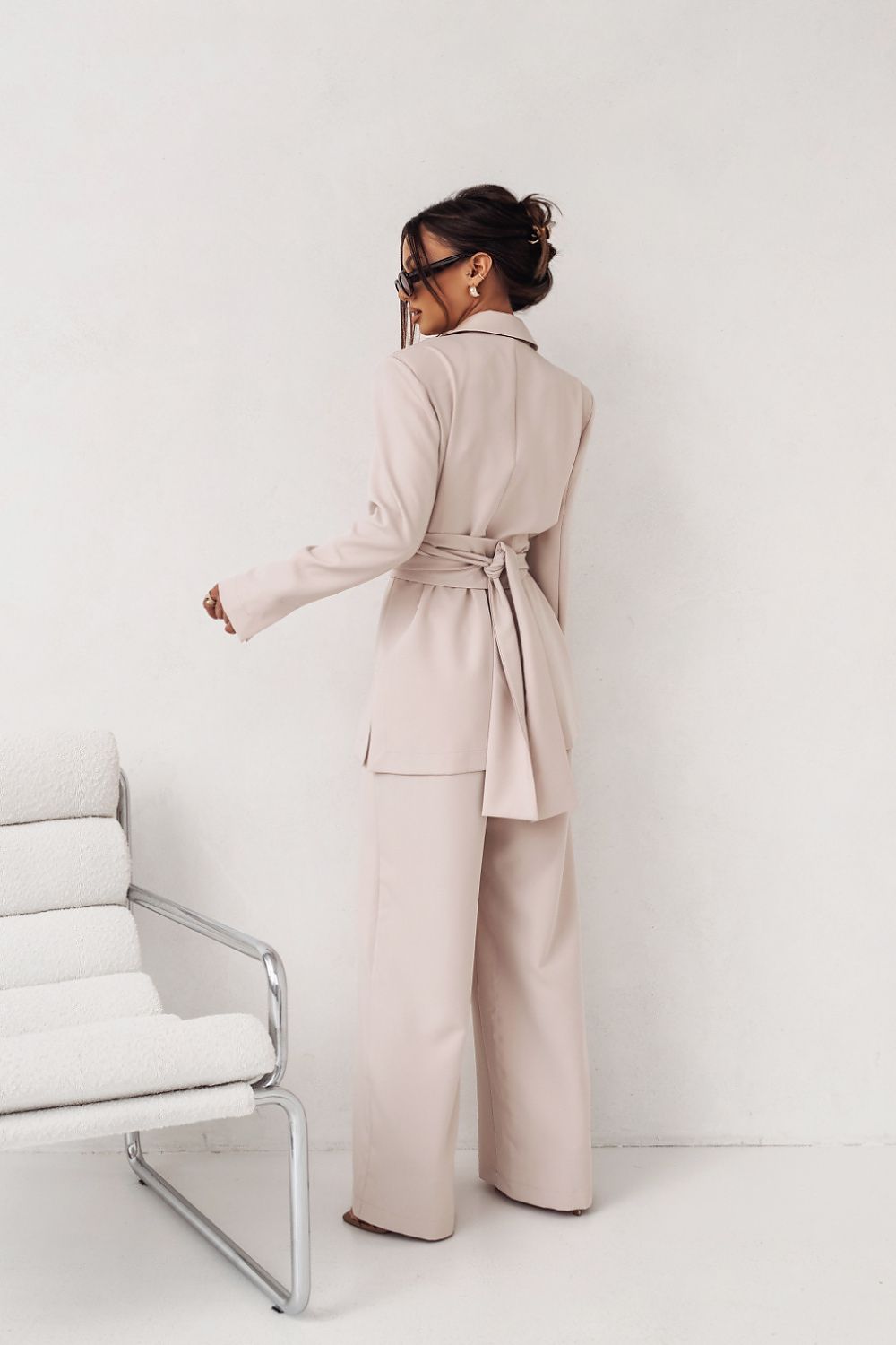 Woman in a beige suit standing in a minimalistic room with a white chair.