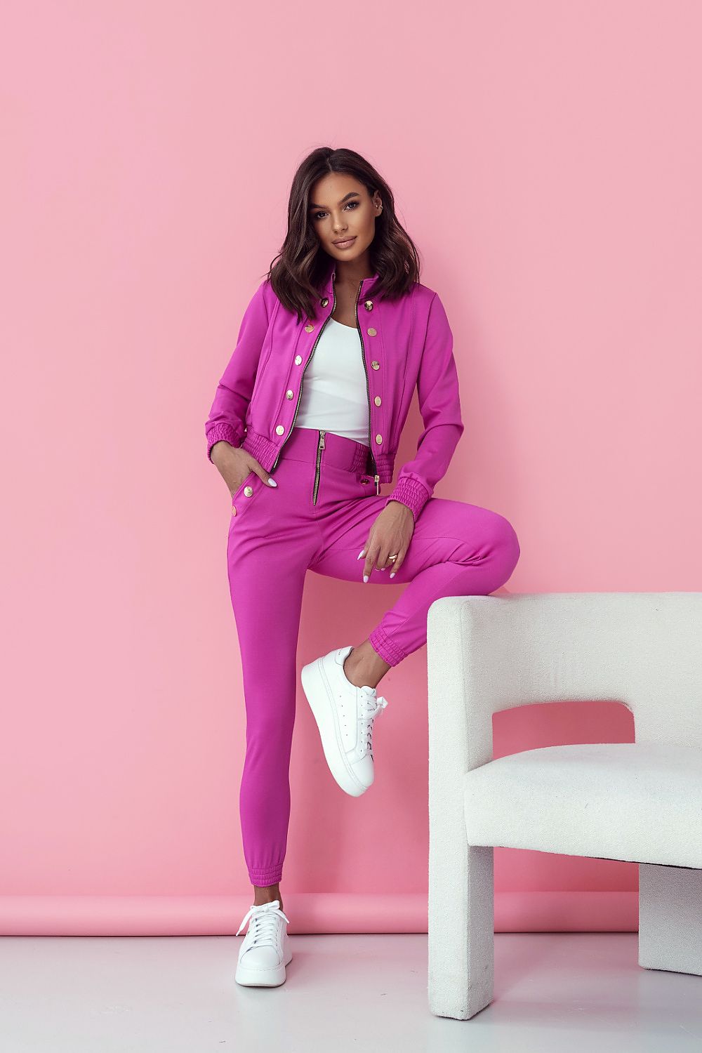 Woman in a pink tracksuit sitting on a white chair against a pink background