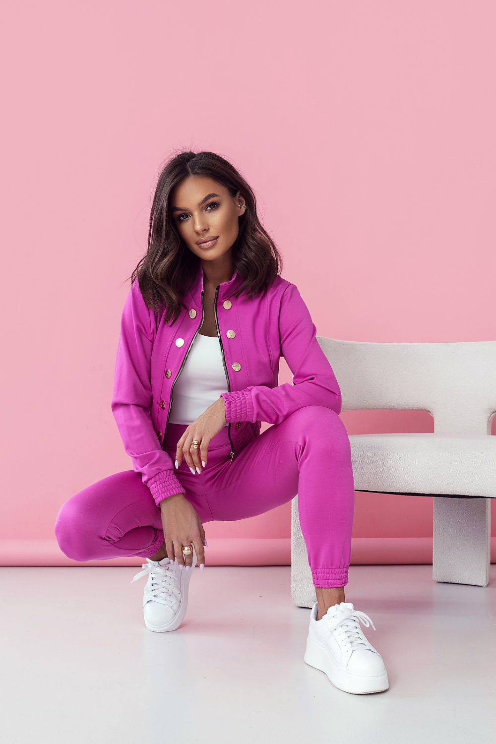 Woman in a bright pink tracksuit sitting on a white chair against a pink background