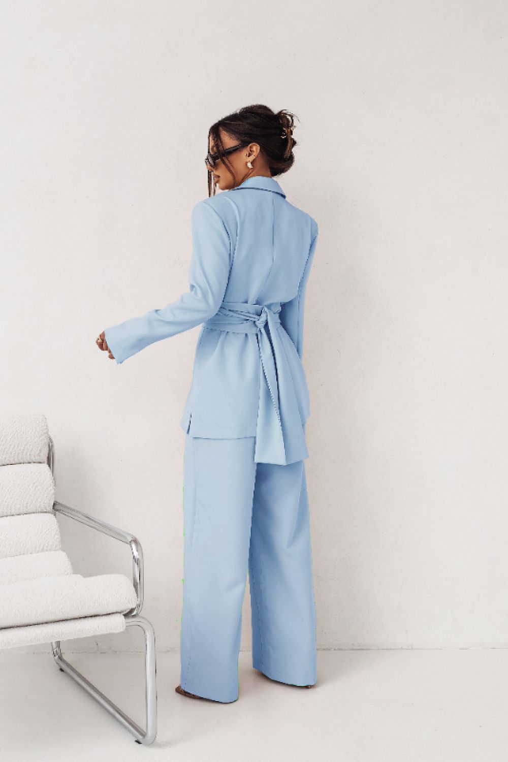 Person wearing a light blue suit standing in a minimalistic room with a white chair.