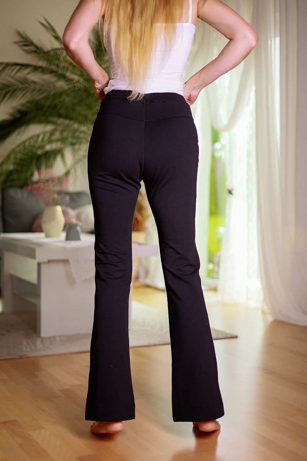 Person wearing black bell-bottom pants in a room with plants and furniture.