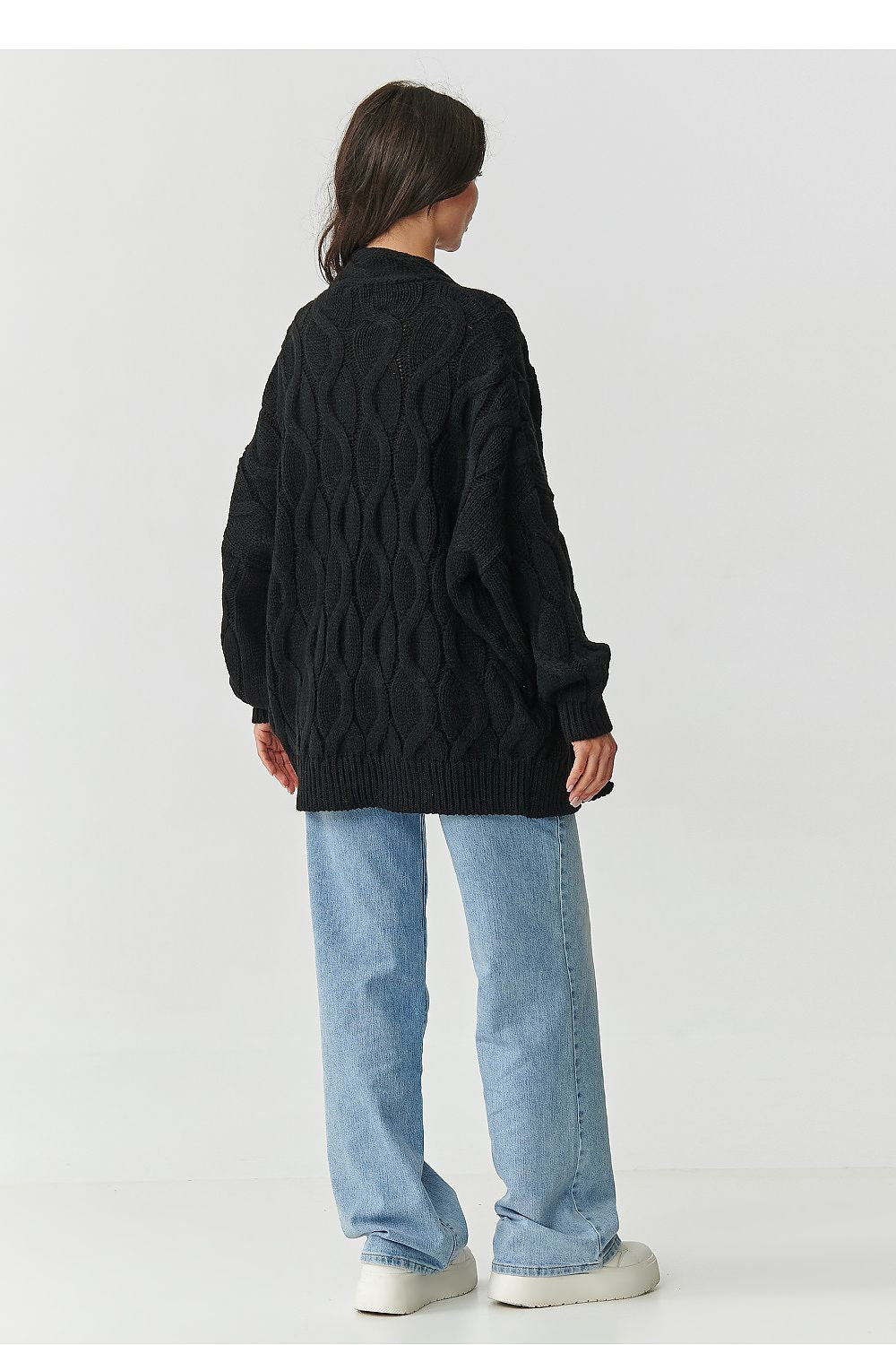 Person wearing a black cable knit sweater and light blue jeans on a white background