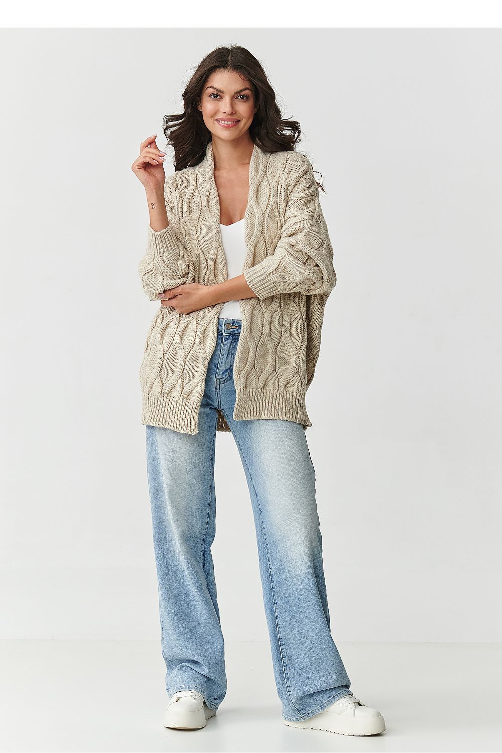 Woman wearing a beige cardigan, white top, and blue jeans on a white background