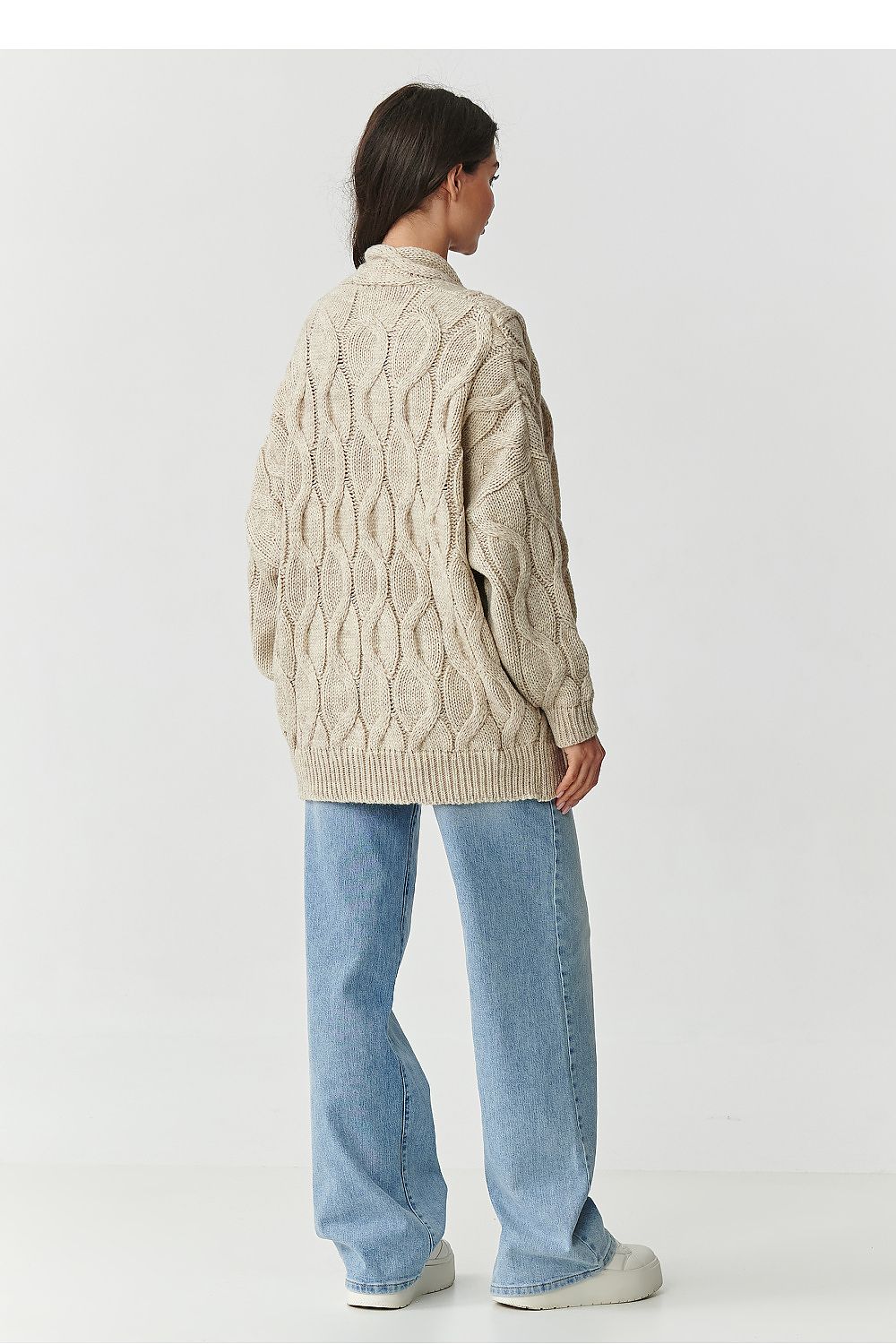 Person wearing a beige cable knit sweater and light blue jeans on a white background