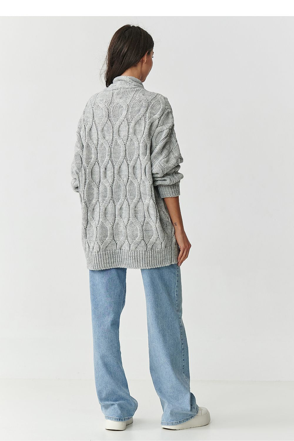 Person wearing a gray cable knit cardigan and light blue jeans on a white background