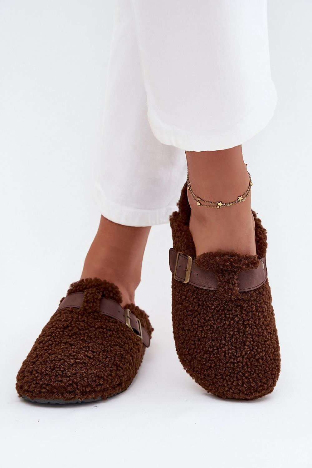 Brown fuzzy slippers worn with white pants on a white background